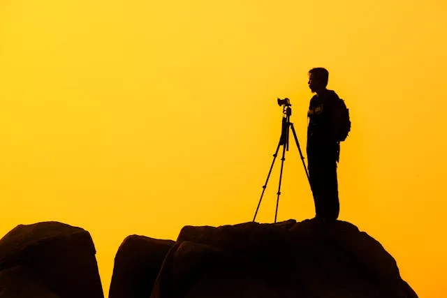 A man stands on a rock with a camera at sunset.