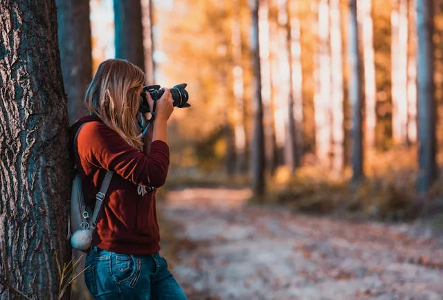 A woman leans on a tree while pointing a camera.