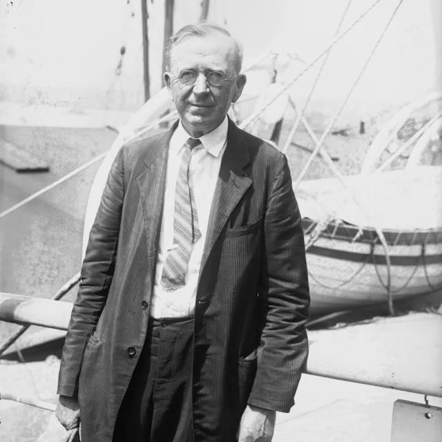 A black and white image of a man in a rumpled suit in front of a boat.