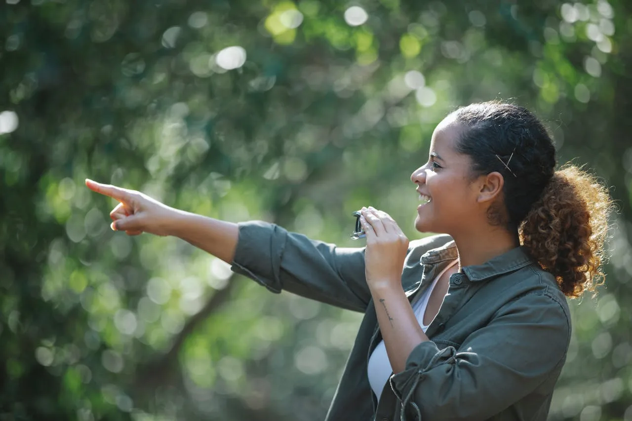 A woman is holding a camera and pointing to some bushes.